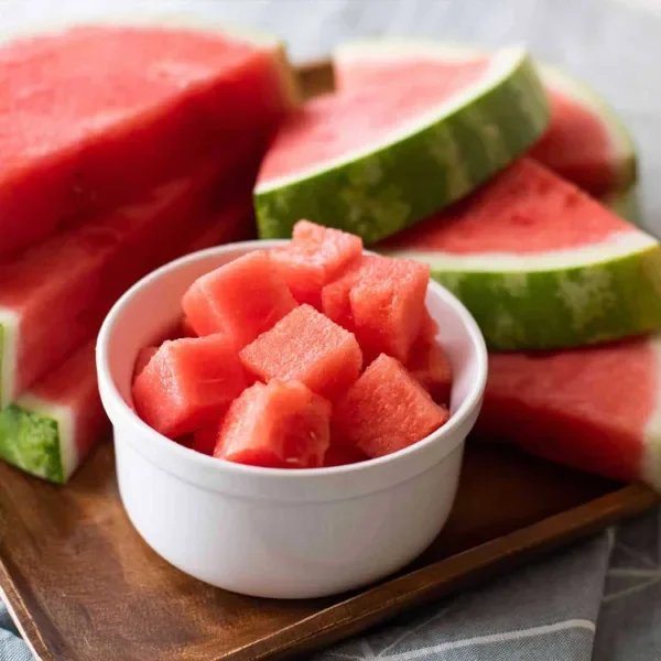 Cut up watermelon in a white bowl, with watermelon slices in the background.