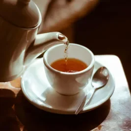 White teacup on a saucer, with hot tea being poured into it.